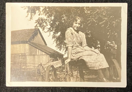 Vintage sepia-toned photograph of a woman sitting on a cart with a barn and trees in the background.