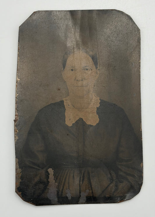 An antique, aged-look portrait photograph of a woman in a dark dress with a light lace collar, mounted on a dark backing with visible wear, tear, and discoloration.