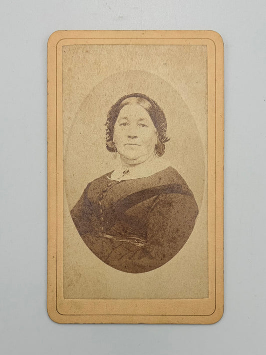 A vintage carte de visite print of a woman, featuring a sepia-toned oval portrait on a tan cardstock with a decorative border, photographed against a neutral light gray background.