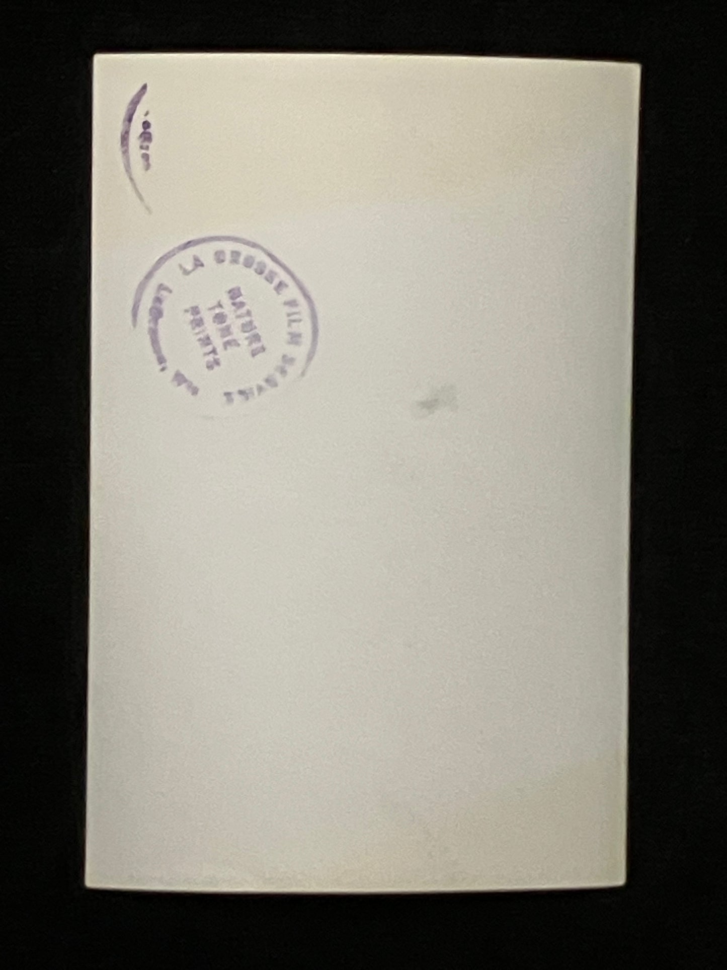 A blank cream-colored photo back with a purple circular stamp on the upper left, displayed against a solid black background.
