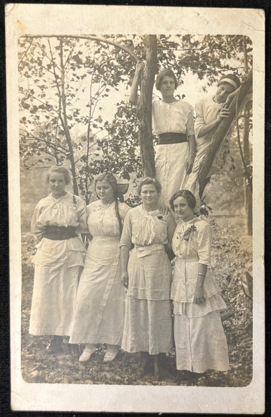 Vintage photograph of six women in white dresses standing outdoors with a tree in the background.
