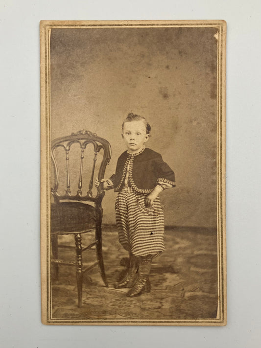 An antique sepia-toned carte de visite photograph showing a young child standing beside a chair against a mottled studio backdrop, with a decorative border around the print.