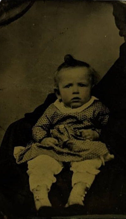 A Victorian era tintype photo of a baby with hand color cheeks, wearing a serious expression.