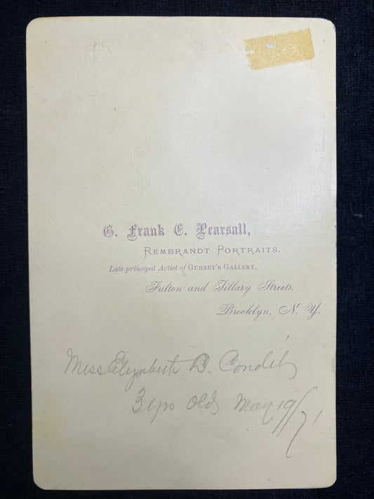 Back of a vintage photograph with text and a yellowed corner on a dark background