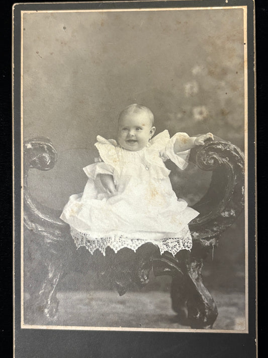 Vintage photograph of a baby in a white dress sitting on a decorative chair.