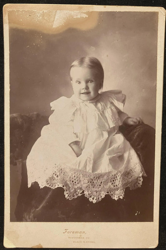 Vintage photograph of a child in a white dress with a lace hem, sitting on a dark surface.