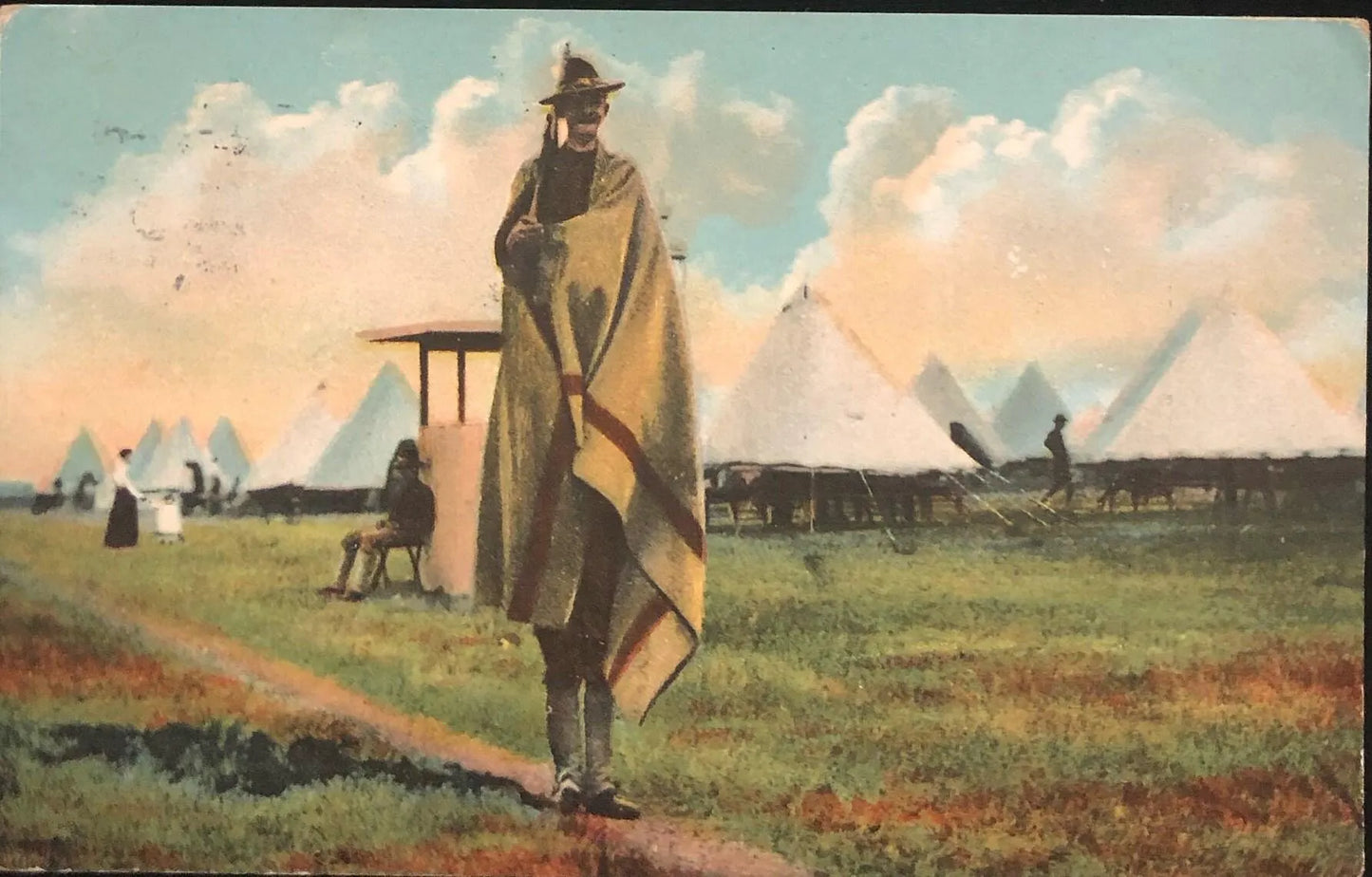 Man in a long coat and hat standing on a grassy field with tents and people in the background