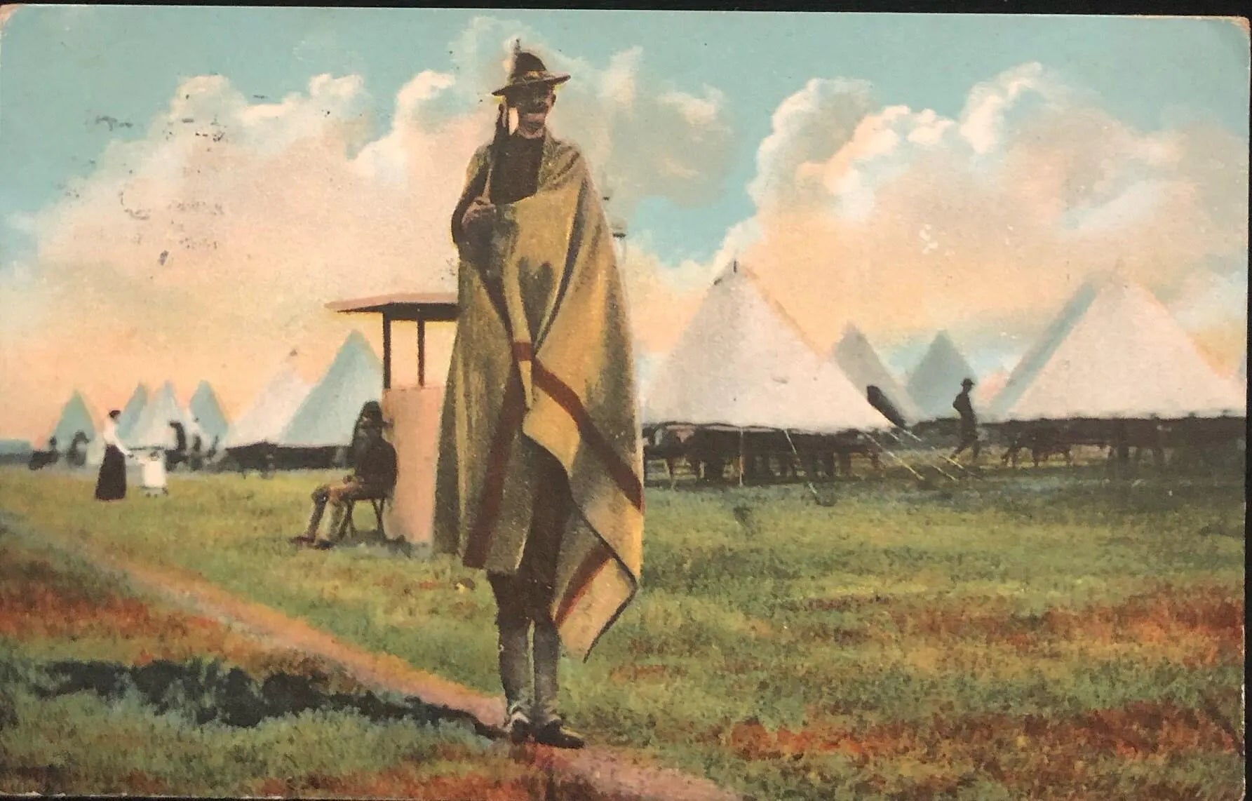 Man in a long coat and hat standing on a grassy field with tents and people in the background