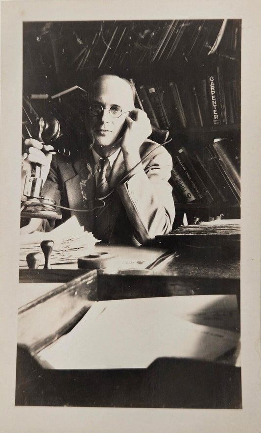 Black and white photo of a man with round glasses on a phone call, taken indoors with natural lighting.
