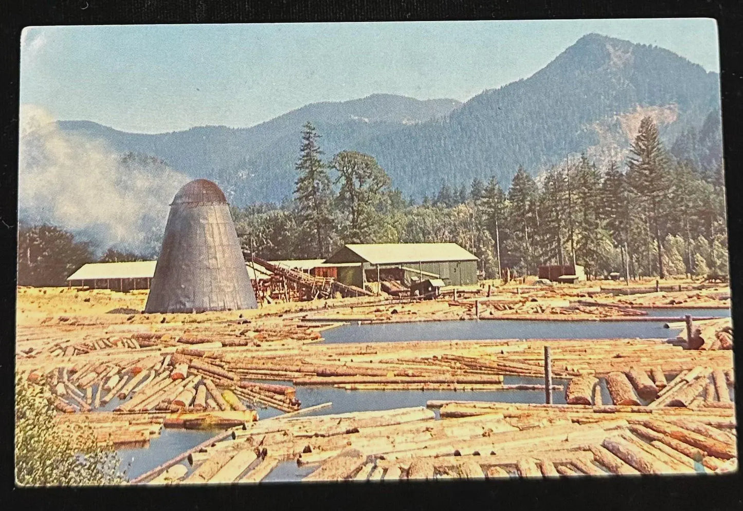 Logging operation with logs, smokestack, and mountains in the background