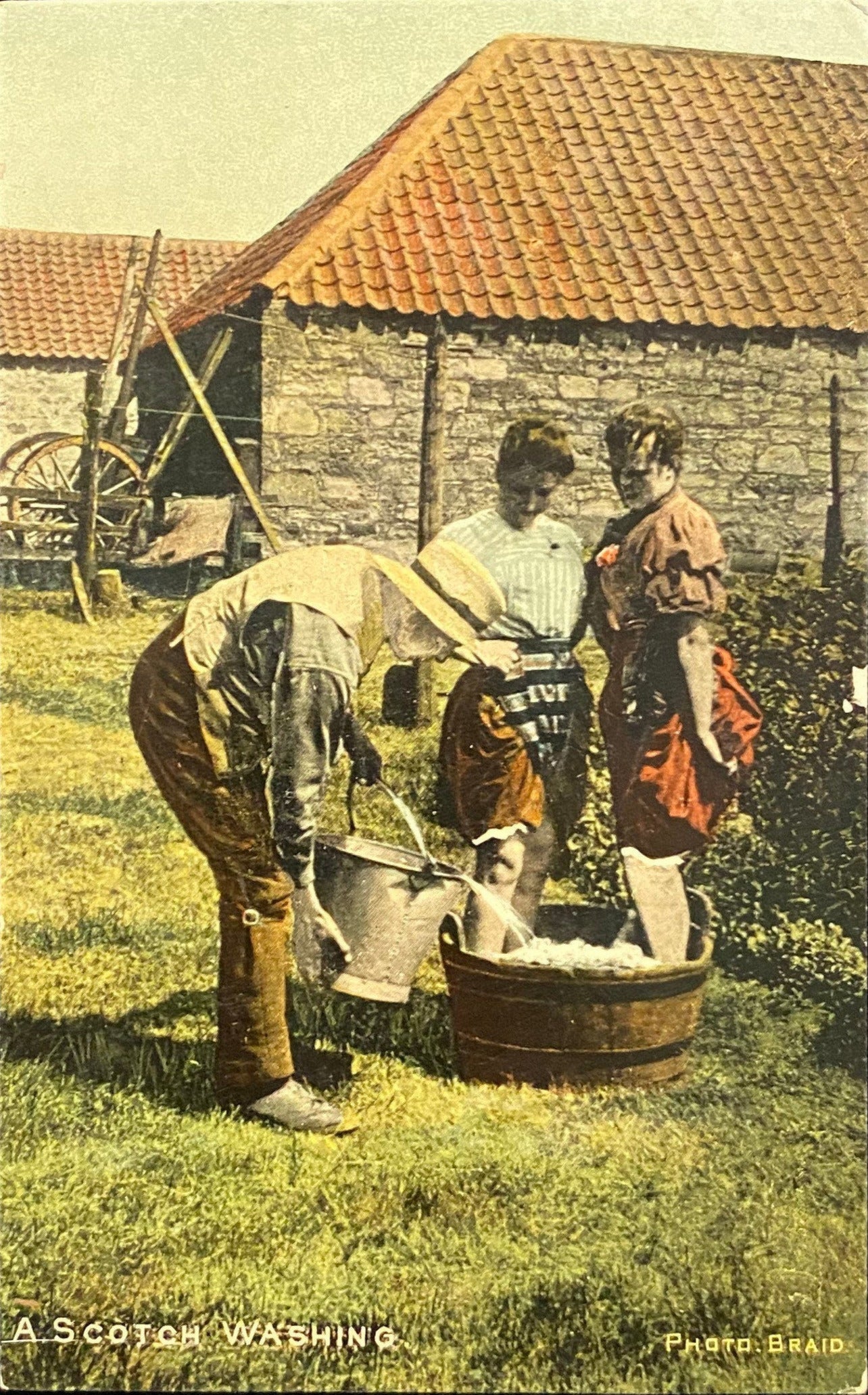 A vintage postcard titled 'A Scotch Washing' showing two individuals engaging in an activity with a large bowl, potentially washing feet, in a rural setting with a thatched roof structure in the background. The postcard includes hand color and has a publication date from the early 20th century.