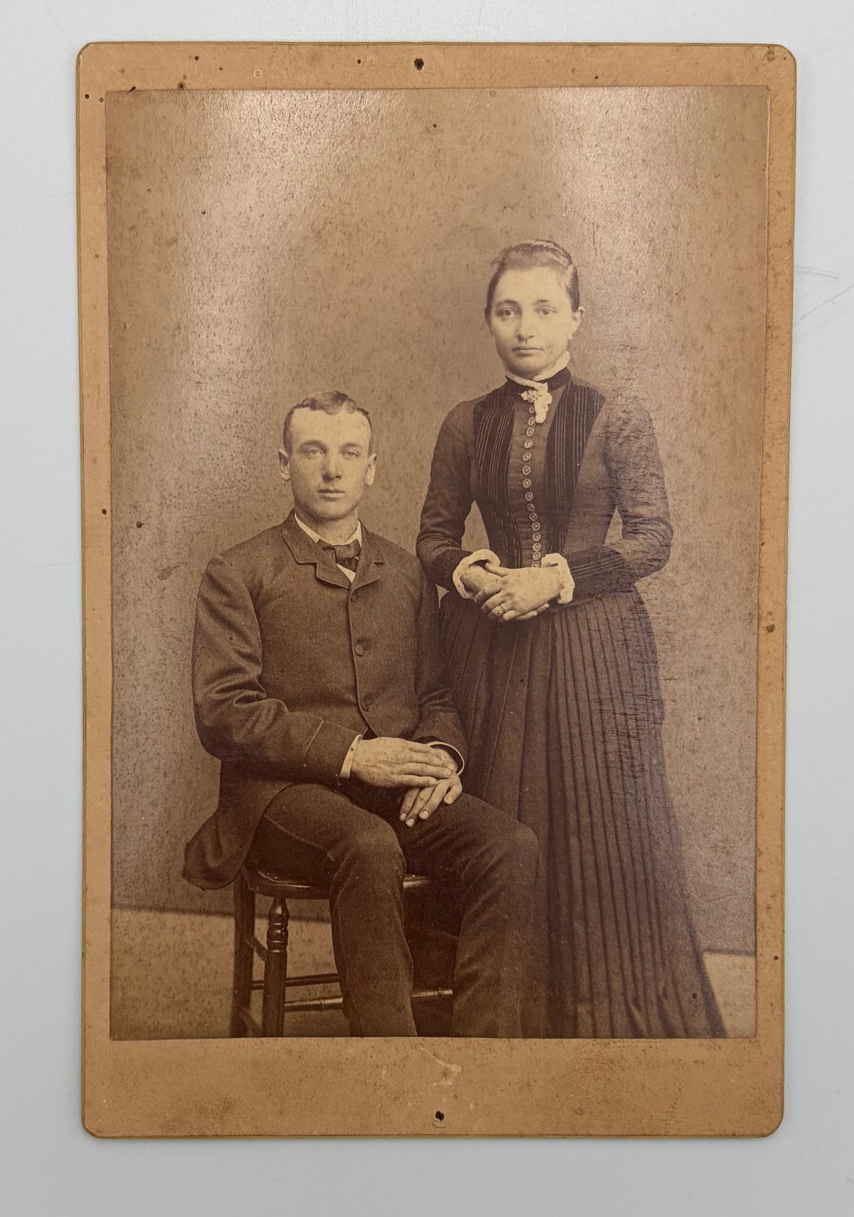 A vintage sepia-toned cabinet card photograph showing a seated man and a standing woman in formal late 19th-century clothing, mounted on a tan paper border against a light gray background.