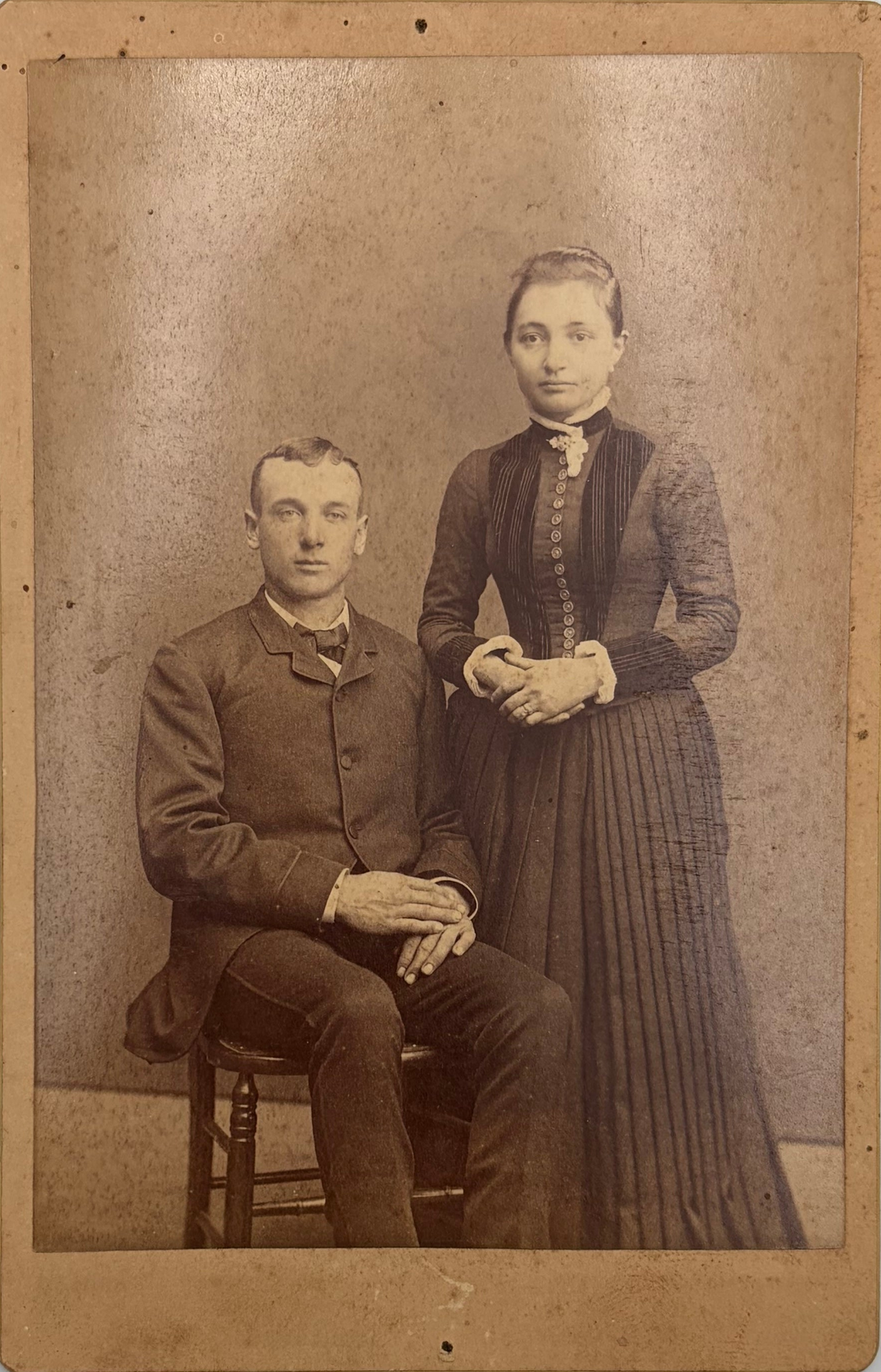 An antique sepia-toned portrait photograph showing a seated man and a standing woman in formal Victorian-era clothing against a mottled studio backdrop.