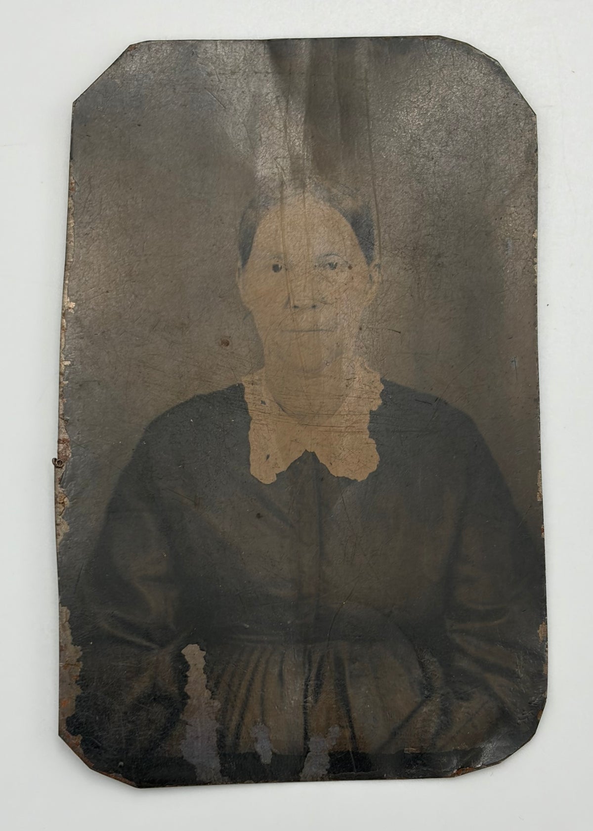 An antique, aged-look portrait photograph of a woman in a dark dress with a light lace collar, mounted on a dark backing with visible wear, tear, and discoloration.