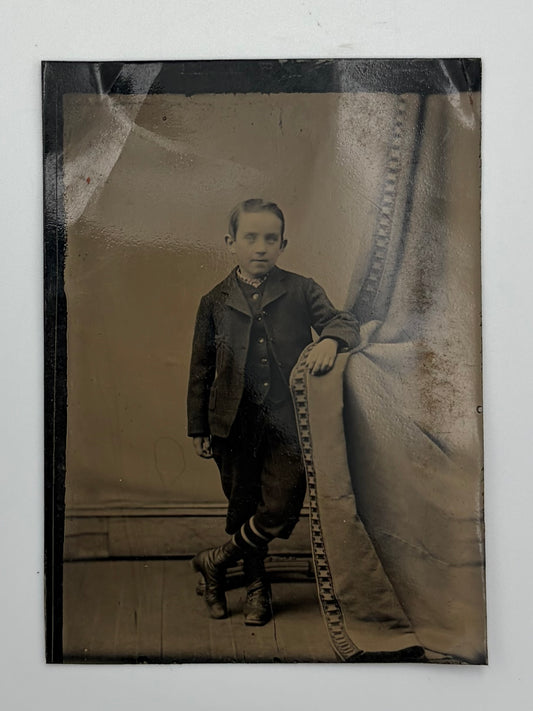 An antique sepia-toned portrait photograph of a young boy in formal clothing, standing beside a draped backdrop. The print shows visible aging, creases, and a dark border, photographed against a light neutral background.