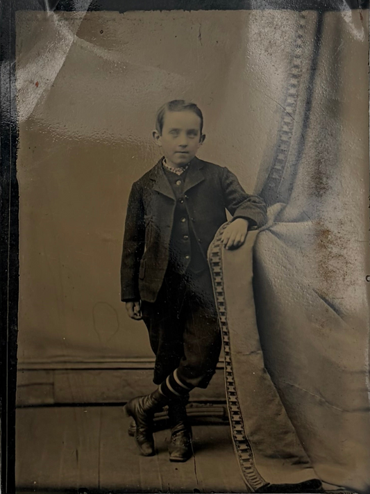 A vintage sepia-toned portrait photograph of a young boy standing in a studio, wearing a dark suit and boots, with a draped backdrop and patterned border trim visible on the right side.