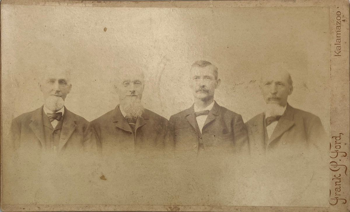 An antique sepia-toned portrait photograph print showing four men in formal suits standing side by side. The print has aged characteristics with visible discoloration, scratches, and a handwritten signature along the right edge reading 'Frank J.