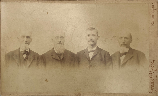 An antique sepia-toned portrait photograph print showing four men in formal suits standing side by side. The print has aged characteristics with visible discoloration, scratches, and a handwritten signature along the right edge reading 'Frank J.