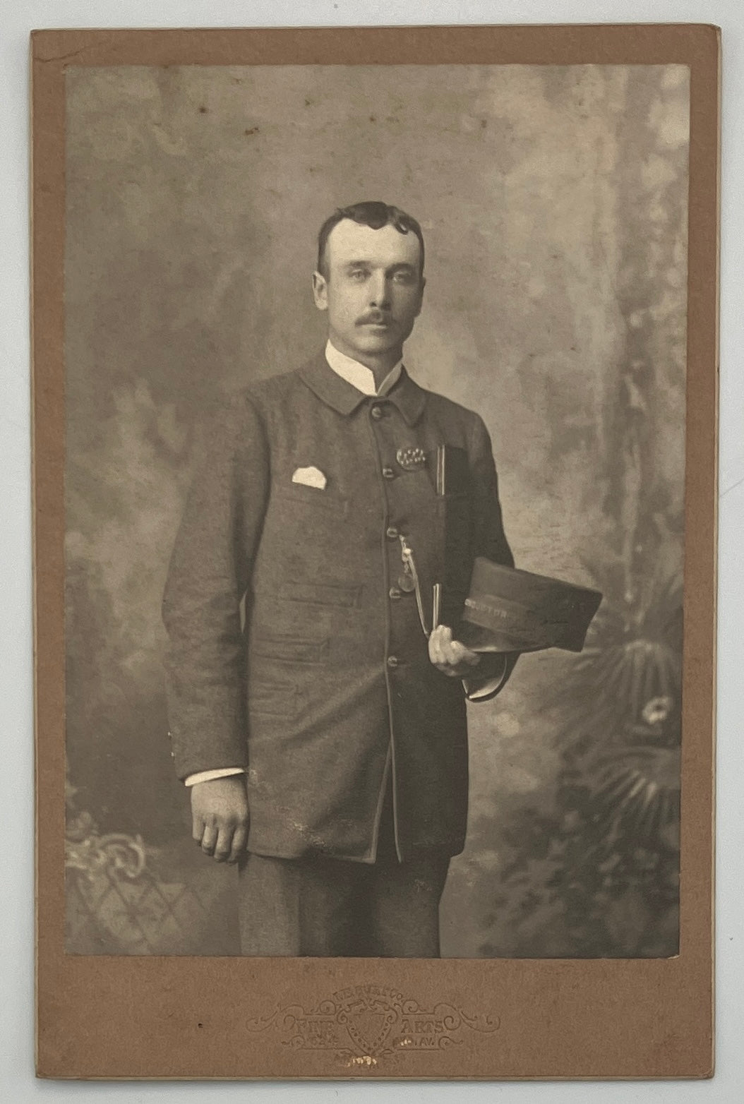 A vintage sepia-toned portrait photograph print of a man in a formal suit holding a hat, mounted on a brown paper border with a mottled studio backdrop.