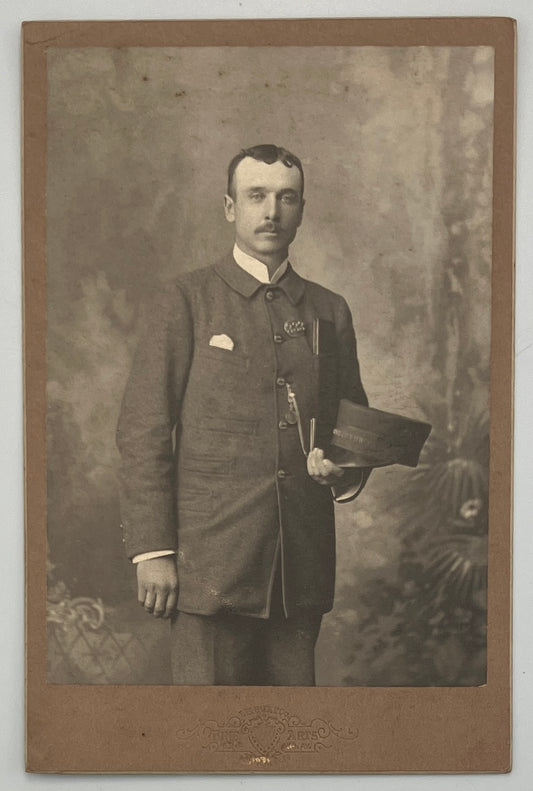 A vintage sepia-toned portrait photograph print of a man in a formal suit holding a hat, mounted on a brown paper border with a mottled studio backdrop.