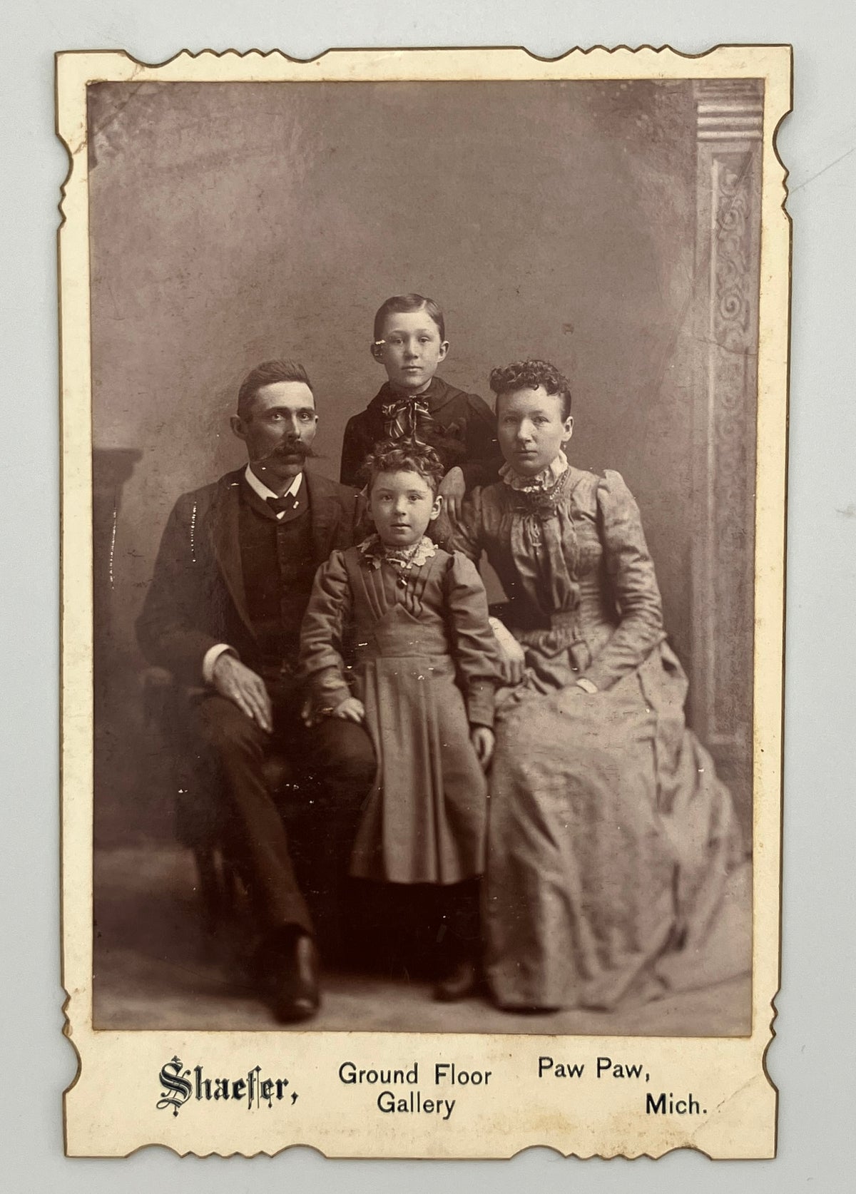 An antique sepia-toned family portrait print with a decorative border and printed text at the bottom reading 'Shaefer, Ground Floor Paw Paw, Mich.' The photo shows four people posed indoors against a studio backdrop.