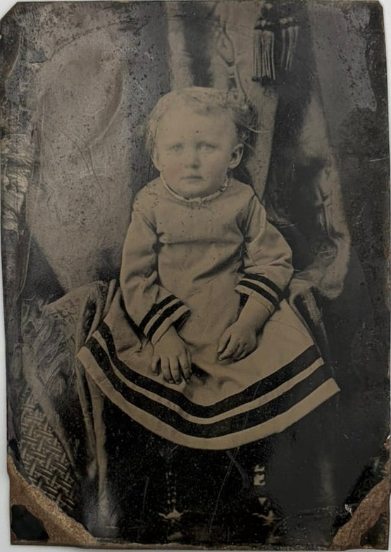 An antique sepia-toned portrait of a young child seated in a dress with dark stripes on the sleeves and hem, photographed against a dark draped backdrop.