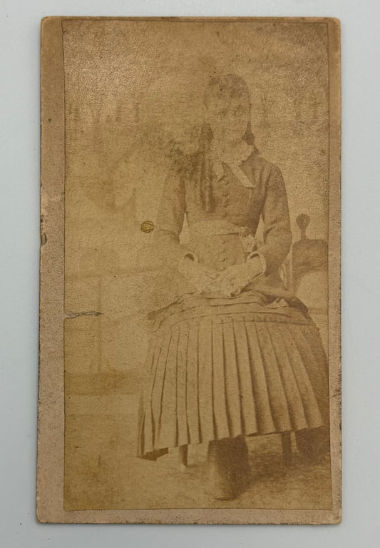 An antique sepia-toned cabinet card photograph showing a seated woman in a long pleated skirt and dark dress, mounted on a tan cardboard border against a light gray background.