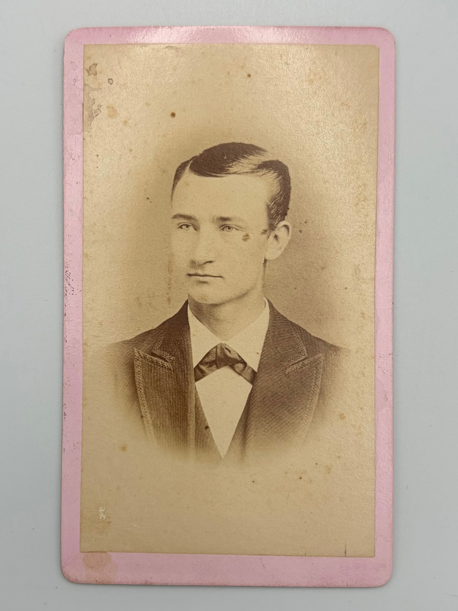 An antique carte de visite portrait of a young man in a dark suit and bow tie, printed on aged beige paper with a pink border, shown against a neutral gray background.