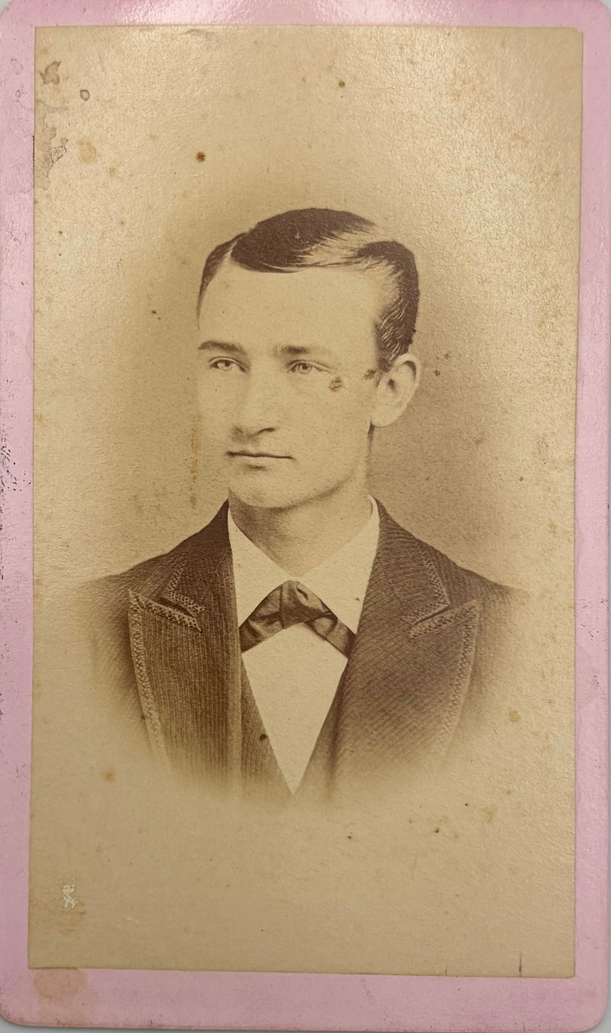 Vintage sepia-toned portrait photograph print of a young man in a suit and bow tie, mounted on a pink card border with age-related discoloration and stains.