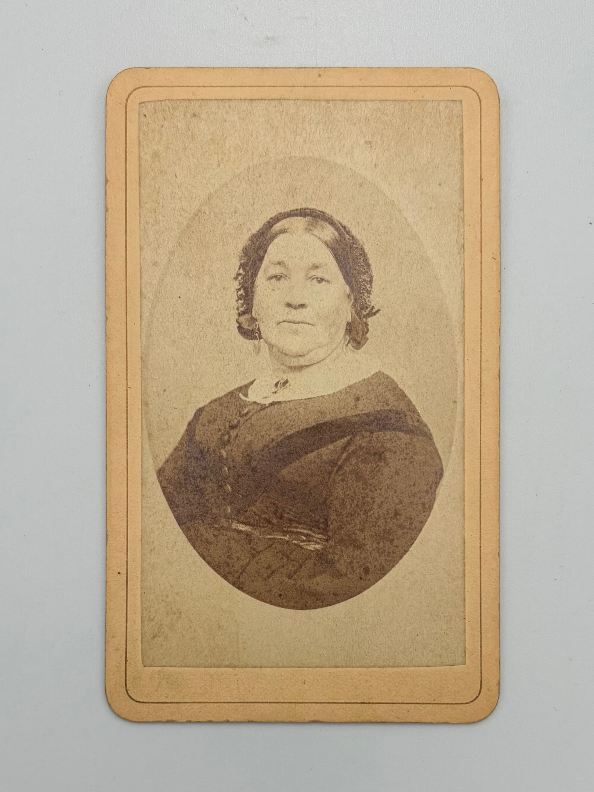 A vintage carte de visite print of a woman, featuring a sepia-toned oval portrait on a tan cardstock with a decorative border, photographed against a neutral light gray background.
