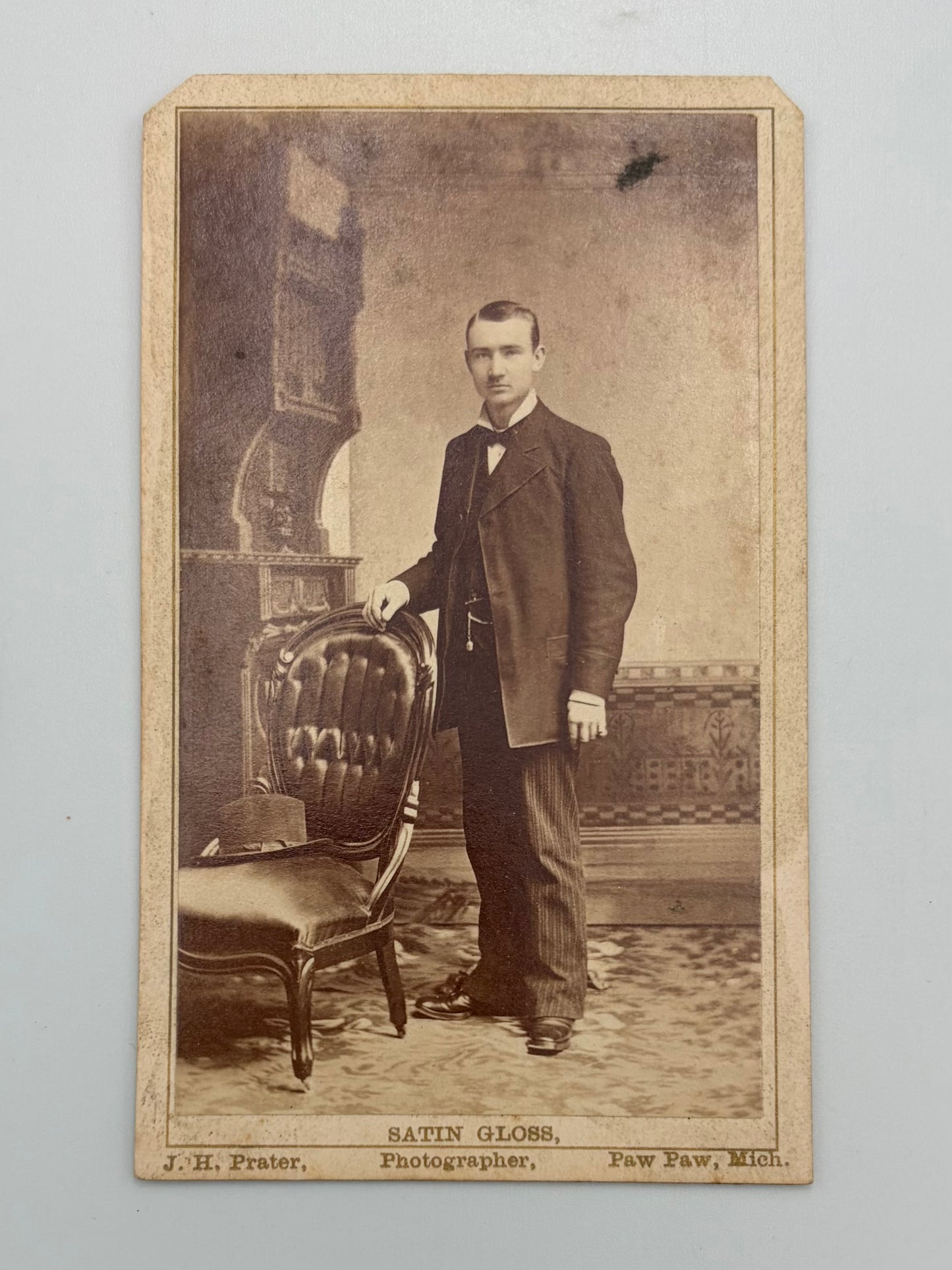 A vintage photograph featuring a man in a dark suit standing beside an upholstered chair, with printed text at the bottom reading 'SATIN GLOSS' and photographer details.