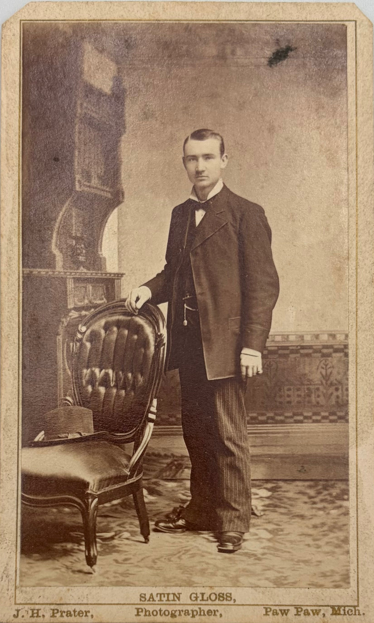 Vintage sepia-toned portrait photograph of a man in formal attire standing beside an upholstered chair, with printed text at the bottom reading 'SATIN GLOSS' and photographer credits.