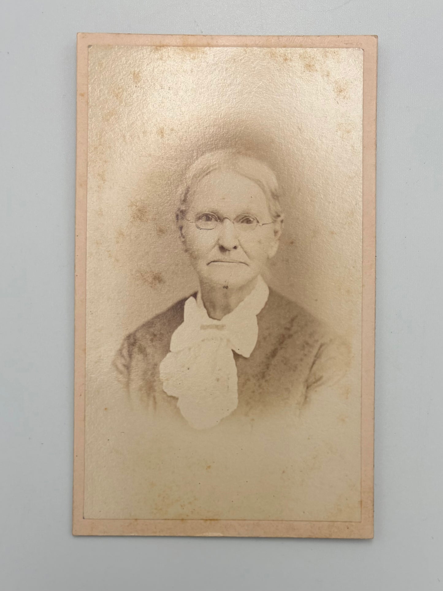 A vintage sepia-toned photograph mounted on a beige card border, showing an elderly woman with glasses and a bow against a plain light gray background.