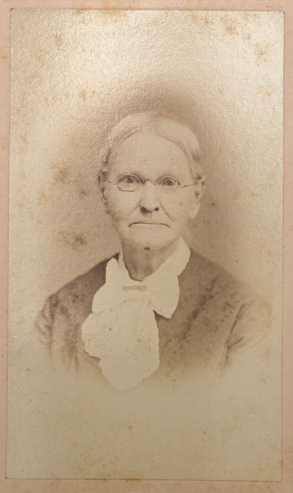 An antique sepia-toned photograph of an elderly woman with glasses, wearing a high white collar, printed on aged paper with a warm beige border.
