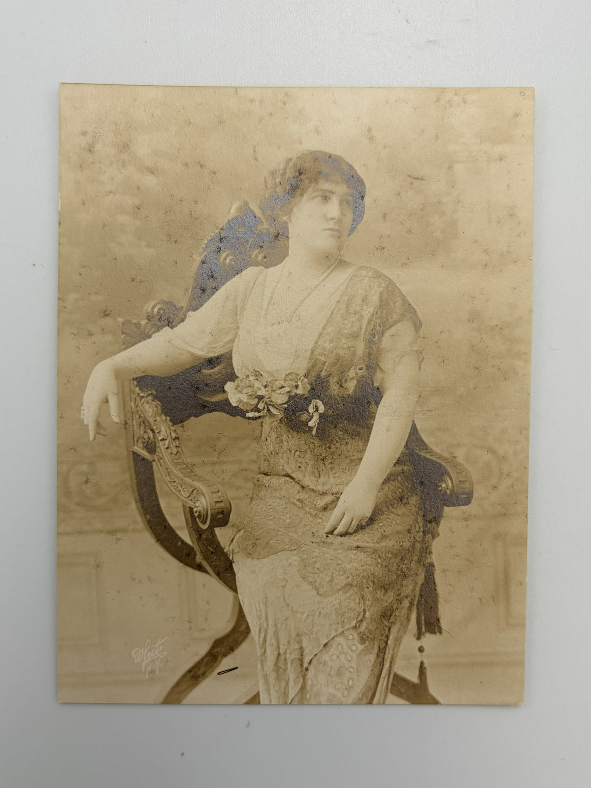 An antique sepia-toned portrait photograph of a woman seated in an ornate chair, shown against a light neutral background.