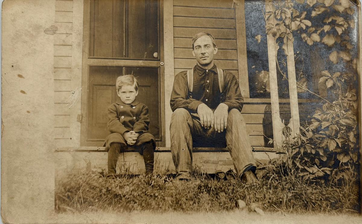 A vintage sepia-toned portrait photograph print showing a seated man and child in front of a house porch, photographed against a light neutral background.