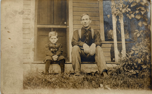 A vintage sepia-toned portrait photograph print showing a seated man and child in front of a house porch, photographed against a light neutral background.