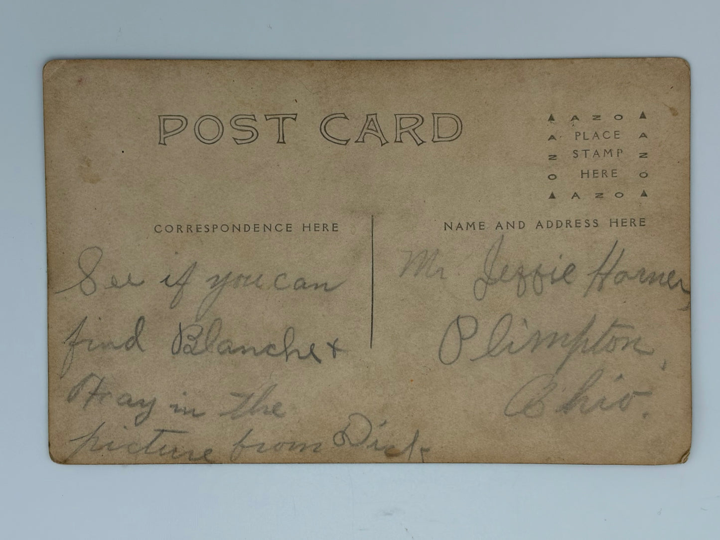 A vintage postcard with a warm tan paper background, featuring printed black text on the top and handwritten cursive writing on both sides. The postcard is photographed flat against a light blue background.