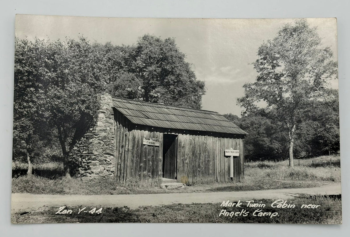 A vintage black and white postcard depicting a rustic wooden cabin surrounded by trees