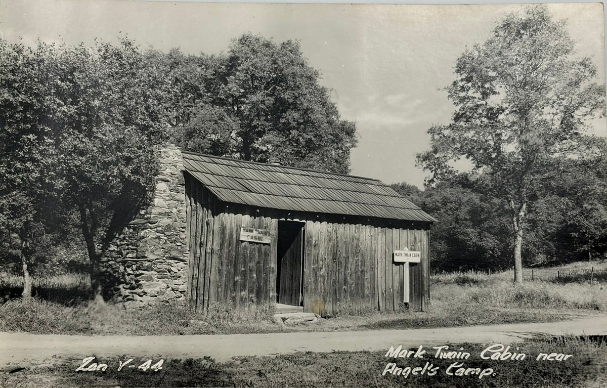 Black and white vintage postcard showing a rustic wooden cabin with a stone chimney, surrounded by trees and open grassy land. Handwritten text appears along the bottom edge.
