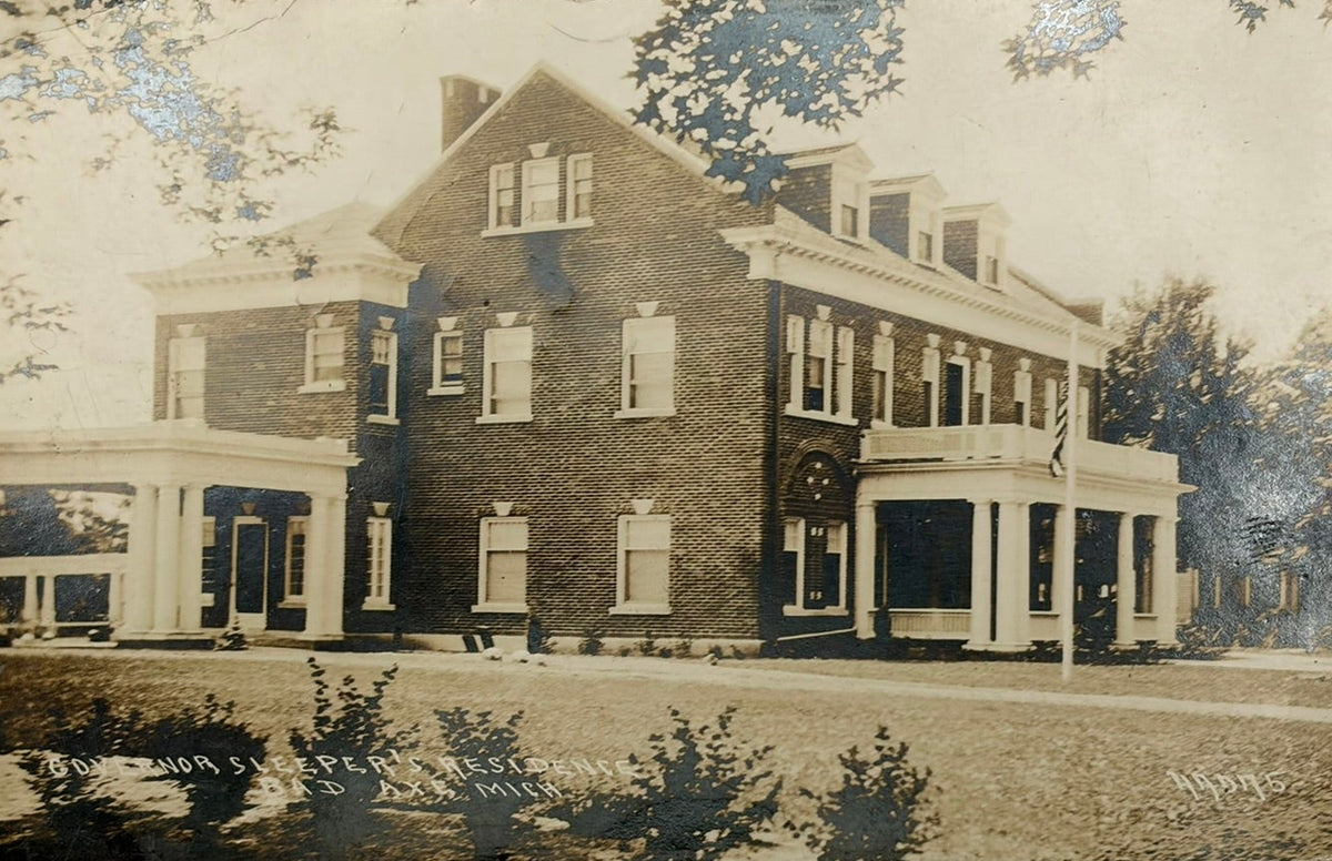 A vintage postcard showing a large brick house with a front porch, displayed on a light gray surface with a coin and ruler for scale.
