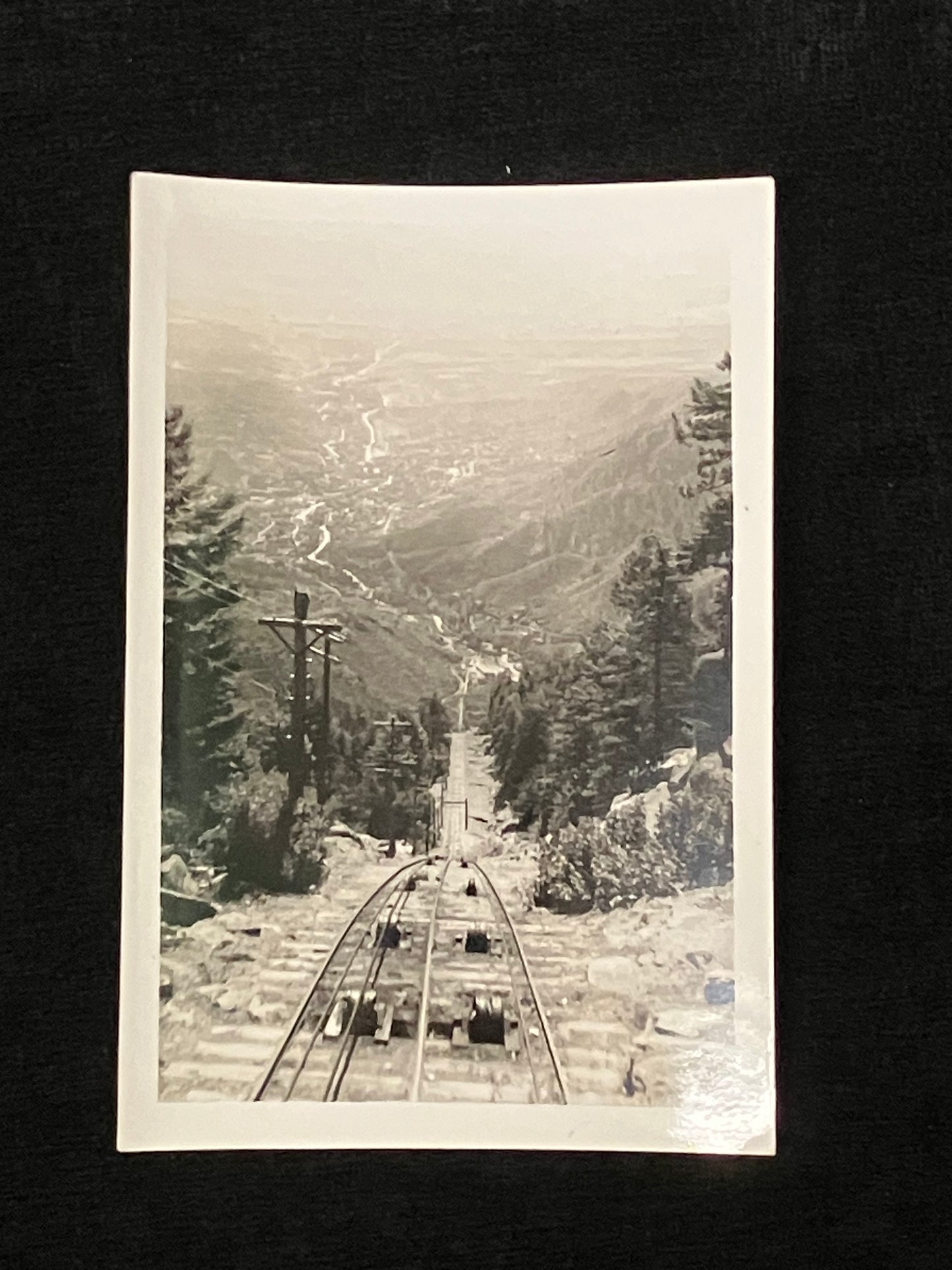 A vintage black and white photograph print depicting a steep railway track leading down a mountain into a valley, surrounded by pine trees, shown against a dark background.
