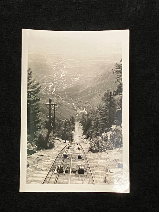 A vintage black and white photograph print depicting a steep railway track leading down a mountain into a valley, surrounded by pine trees, shown against a dark background.