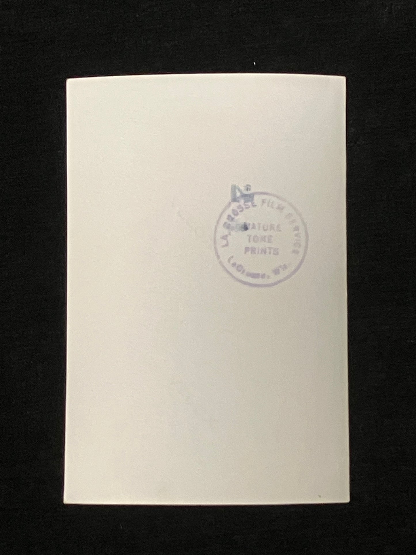 A blank off-white rectangular photo back with a faint circular blue stamp near the top right, photographed against a solid black background.