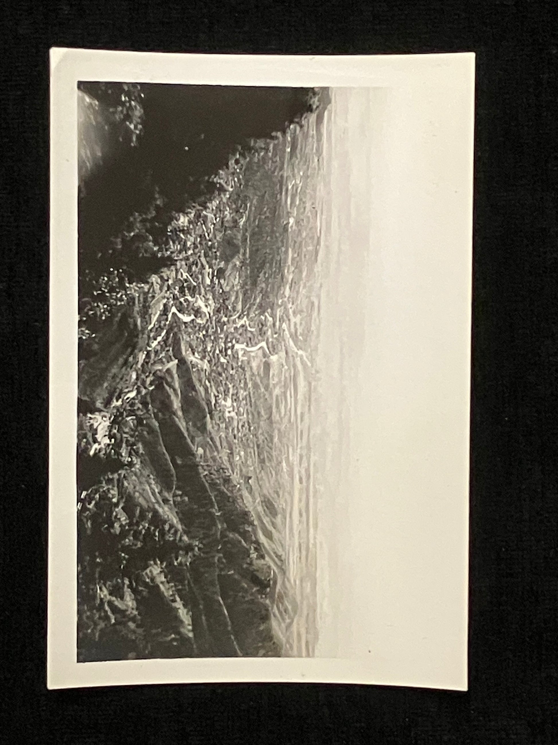 A black and white photographic print or postcard showing a dramatic mountain valley scene, photographed against a black background.