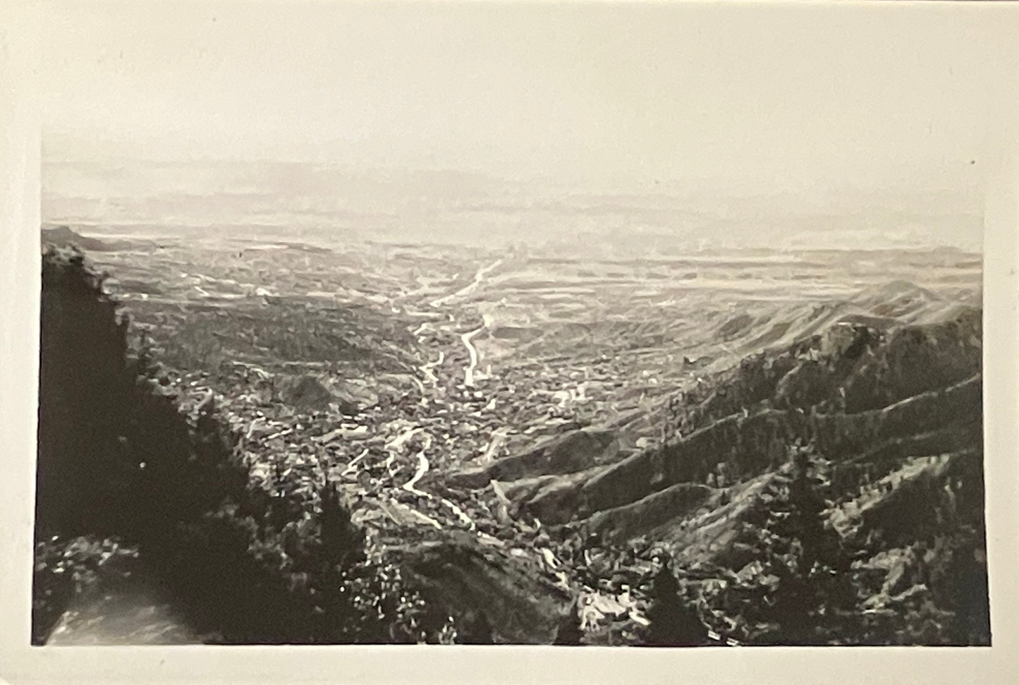 A black and white vintage landscape photograph showing a wide valley with winding roads, hills, and trees in the foreground, presented with a white border.