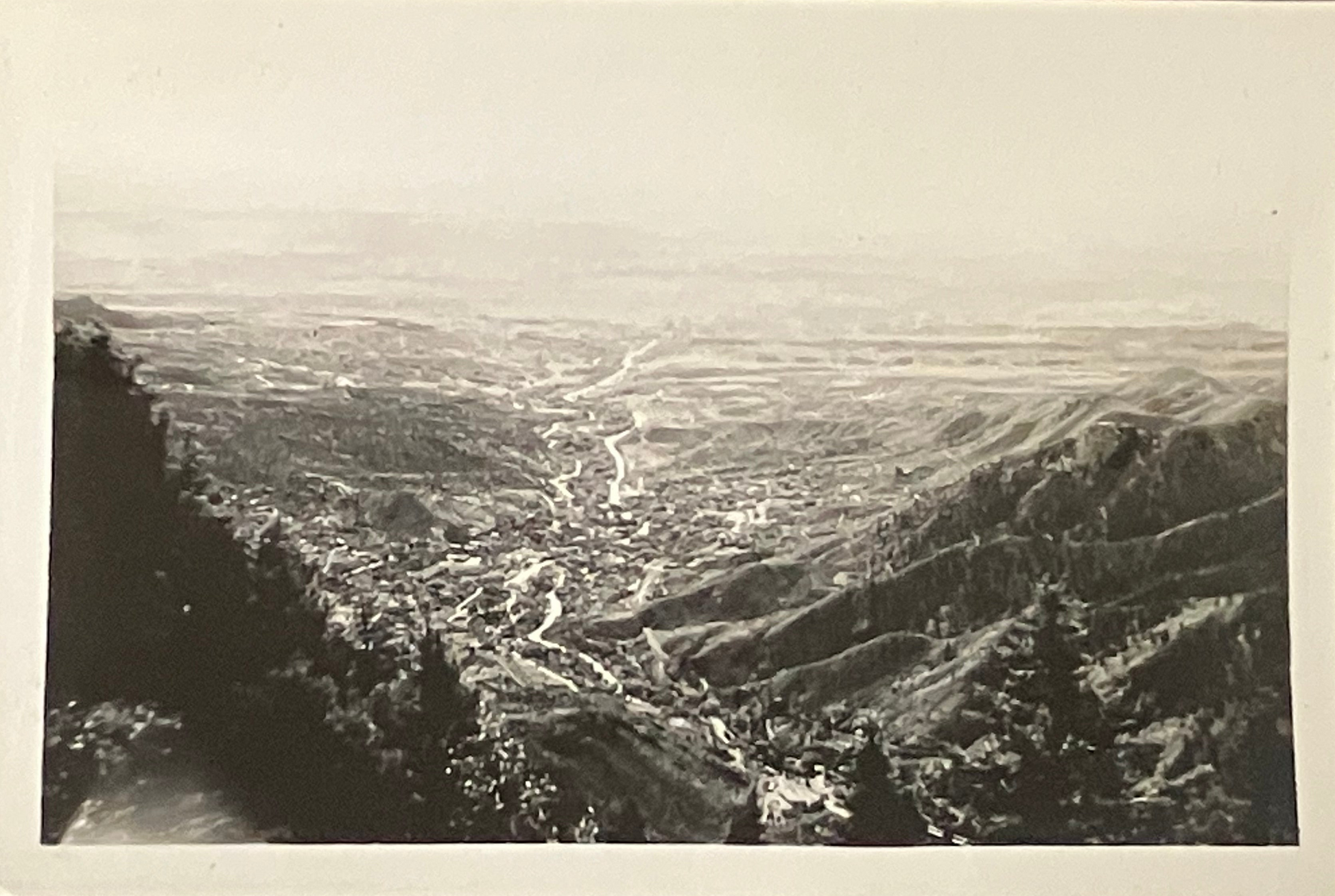 A black and white vintage landscape photograph showing a wide valley with winding roads, hills, and trees in the foreground, presented with a white border.