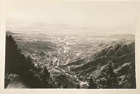 A black and white vintage landscape photograph showing a wide valley with winding roads, hills, and trees in the foreground, presented with a white border.