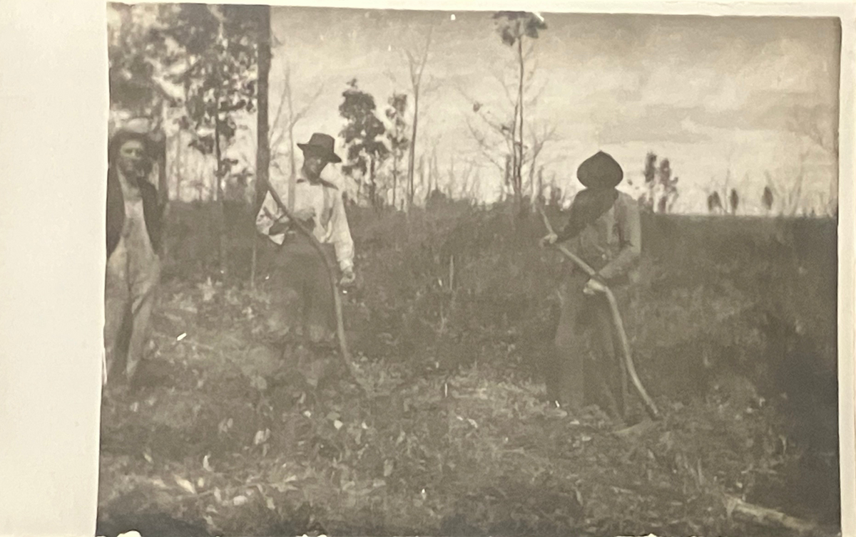 An antique sepia-toned photograph depicting three men working in a field, holding hand tools. The print shows aged paper texture, warm brown and beige tones, and a hazy, vintage aesthetic.