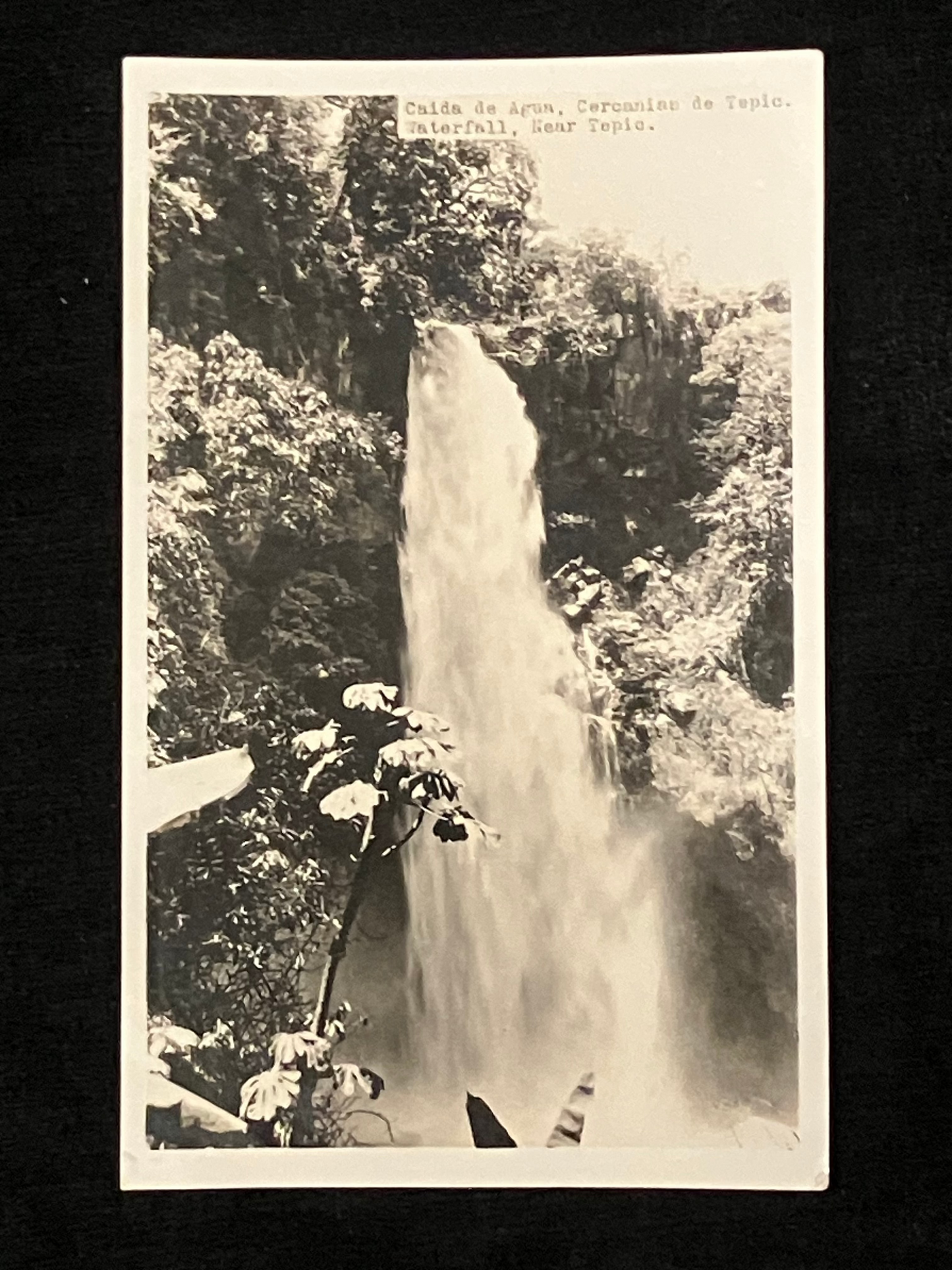 A vintage black and white postcard showing a waterfall cascading down a rocky cliff, with a person standing on a platform to the right. The postcard is photographed against a solid black background.